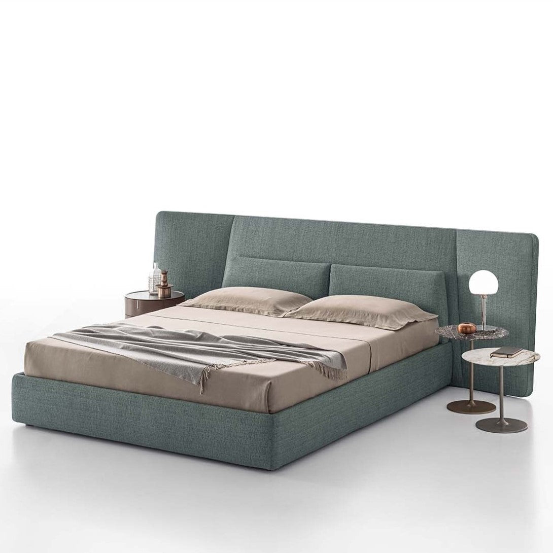 A contemporary Coventry Naked bed with a upholstered headboard, dressed in beige bedding, flanked by a nightstand and a lamp on a round table.