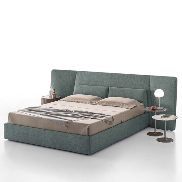 A contemporary Coventry Naked bed with a upholstered headboard, dressed in beige bedding, flanked by a nightstand and a lamp on a round table.